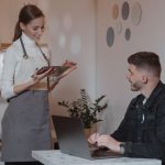A smiling waitress takes an order from a customer on a tablet in a cozy café setting.