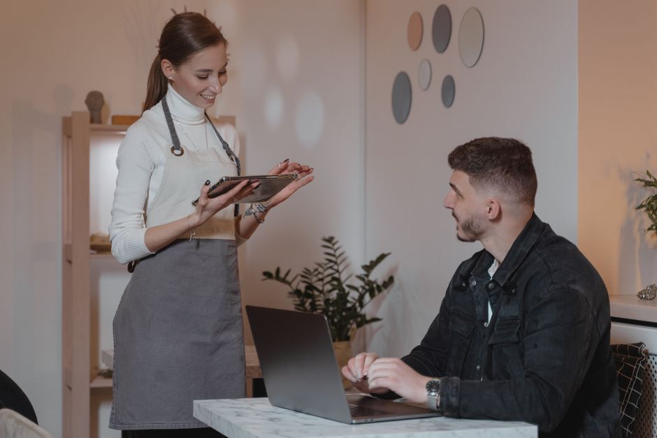 A smiling waitress takes an order from a customer on a tablet in a cozy café setting.